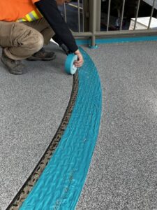 Worker in an orange vest taping a blue seal around a curved concrete edge near metal railings.
