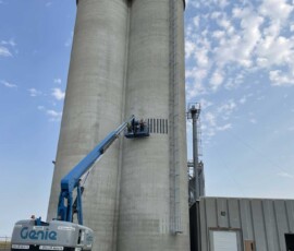 A worker in a boom lift platform cleans a section of a tall concrete grain silo.