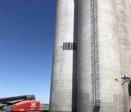 A red boom lift parked next to tall concrete grain silos with an attached metal ladder.