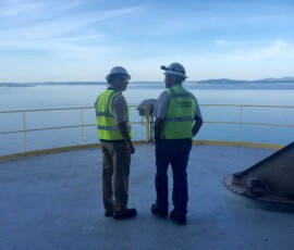 Two workers in hard hats and high-visibility vests stand on an observation deck overlooking a large body of water.